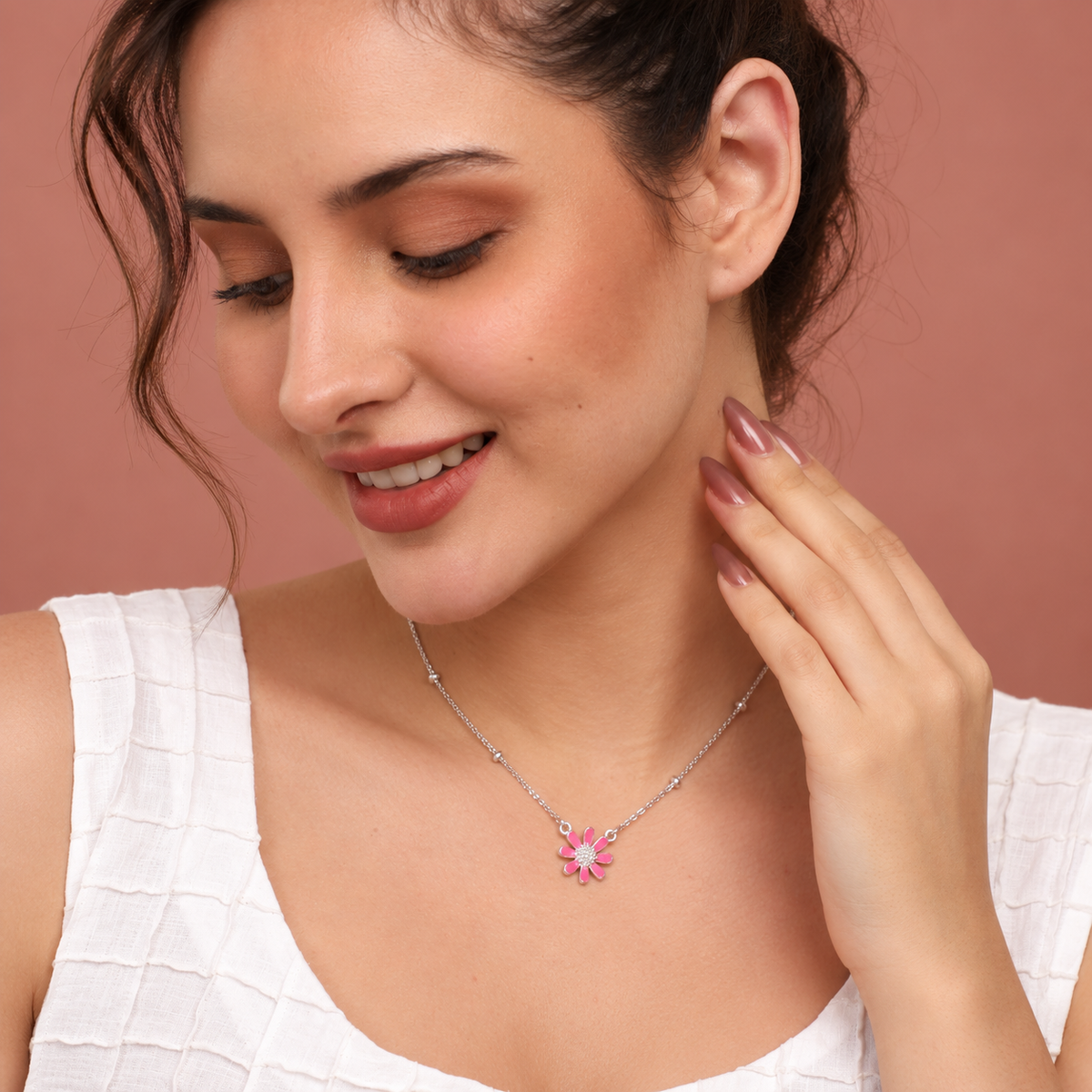 A woman wearing a delicate silver chain necklace with a pink flower-shaped pendant, posing against a solid dusty rose background.