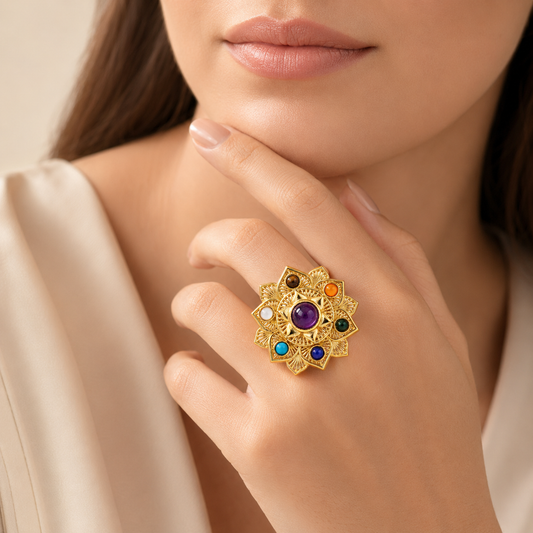 Close-up lifestyle shot of a woman wearing a large, ornate gold ring with multiple colorful gemstones on her finger. She is wearing a cream-colored top, and the background is a soft, neutral beige.

