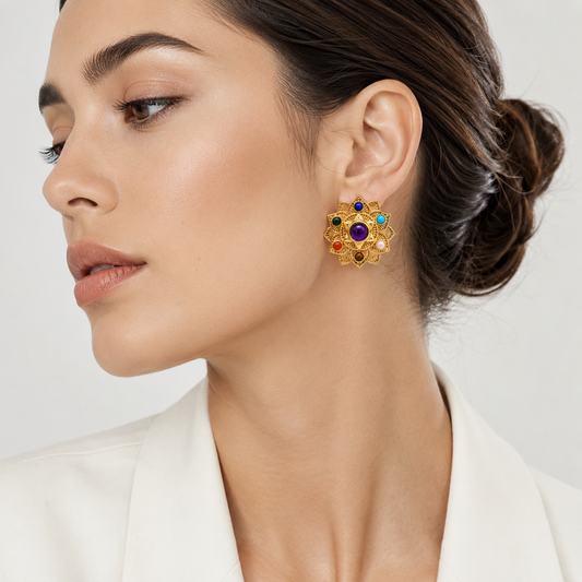 A close-up of a woman wearing a gold-toned floral stud earring with colorful gemstone accents, including a prominent purple center stone.


