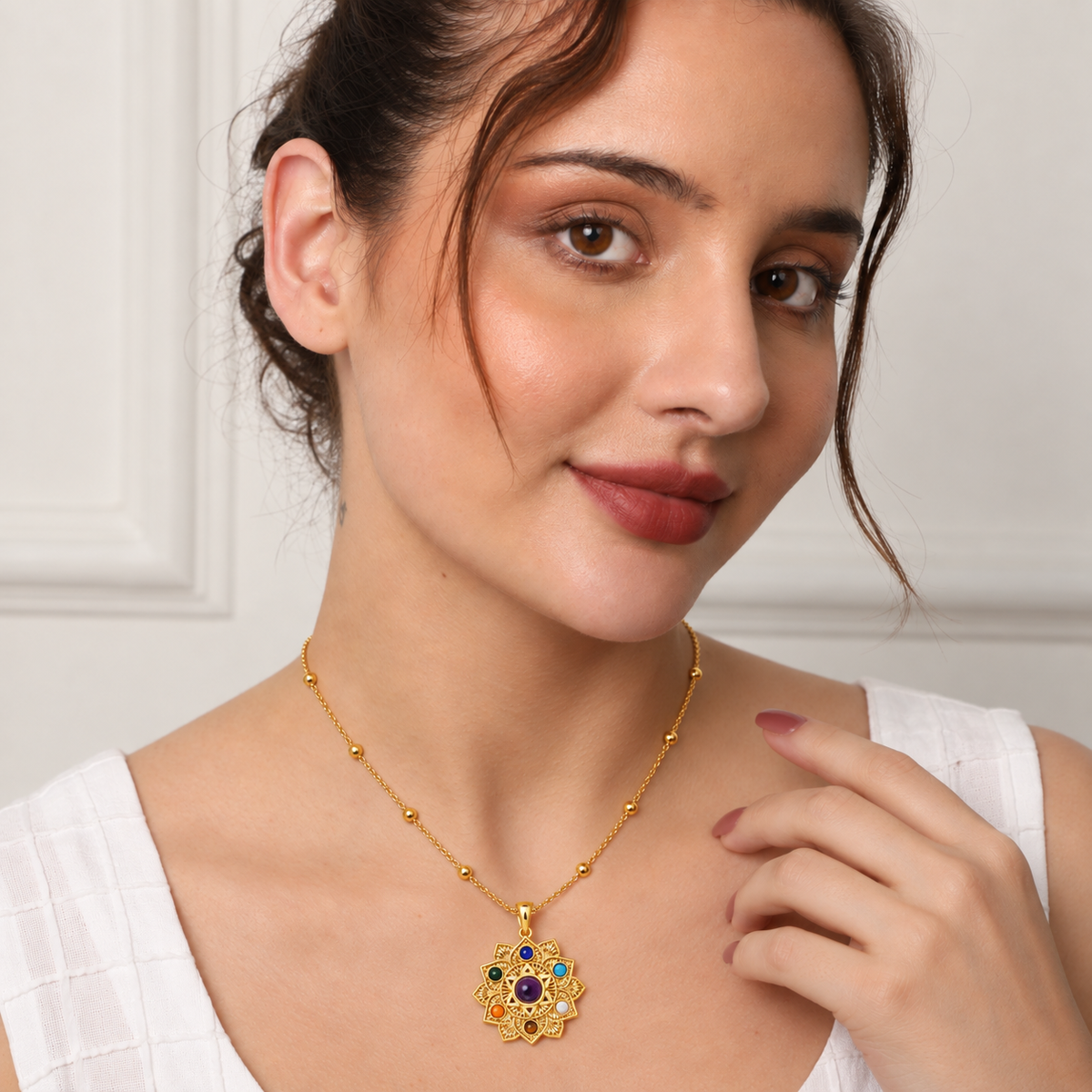 A woman wearing a gold floral pendant necklace with colorful gemstone accents, photographed in a clean indoor setting.


