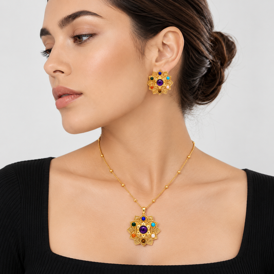 A woman wearing a gold necklace with a matching flower-shaped pendant and matching stud earrings, featuring multicolored gemstone accents, set against a light gray background.

