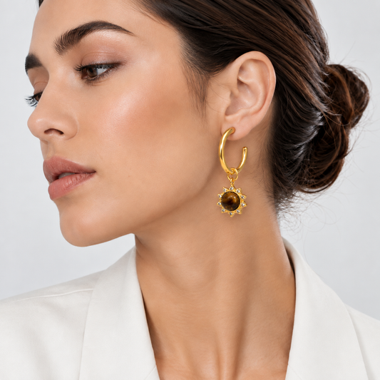 A close-up of a woman wearing a gold huggie hoop earring with a dangling round amber-brown stone and small clear accent stones, paired with a white blazer against a light gray background.

