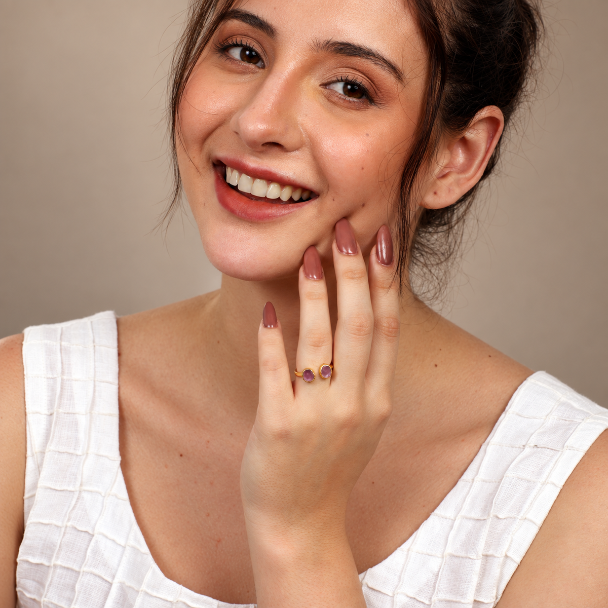 A close-up lifestyle shot of a smiling woman with her hand raised to her face, wearing a delicate gold ring with two small purple gemstones on her ring finger. She is wearing a white textured top against a neutral beige background.


