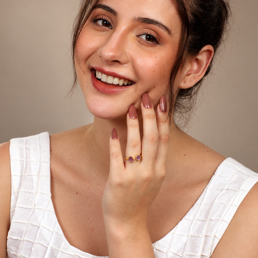 A close-up lifestyle shot of a smiling woman with her hand raised to her face, wearing a delicate gold ring with two small purple gemstones on her ring finger. She is wearing a white textured top against a neutral beige background.

