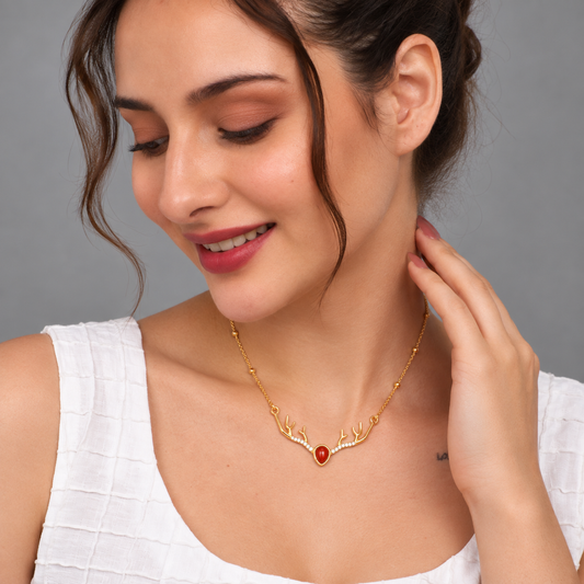 A woman wearing a delicate gold necklace with a heart-shaped antler design and a central red gemstone, shown in a close-up portrait against a neutral gray background.

