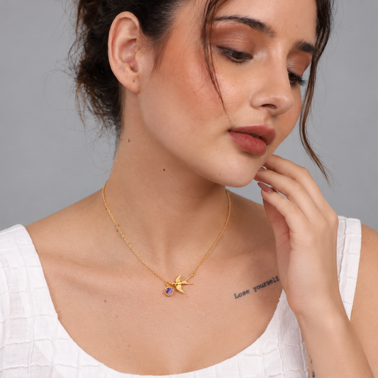 A woman wearing a delicate gold chain necklace with a starfish pendant and a small purple gemstone accent, photographed against a neutral gray background.

