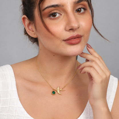 A woman wearing a delicate gold chain necklace with a gold swallow pendant and a small green oval stone, shown in a close-up portrait against a neutral gray background.

