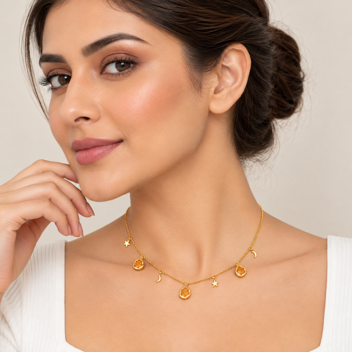 A woman wearing a delicate gold necklace with small celestial charms, including stars, moons, and teardrop pendants, styled with a white top against a soft beige background.


