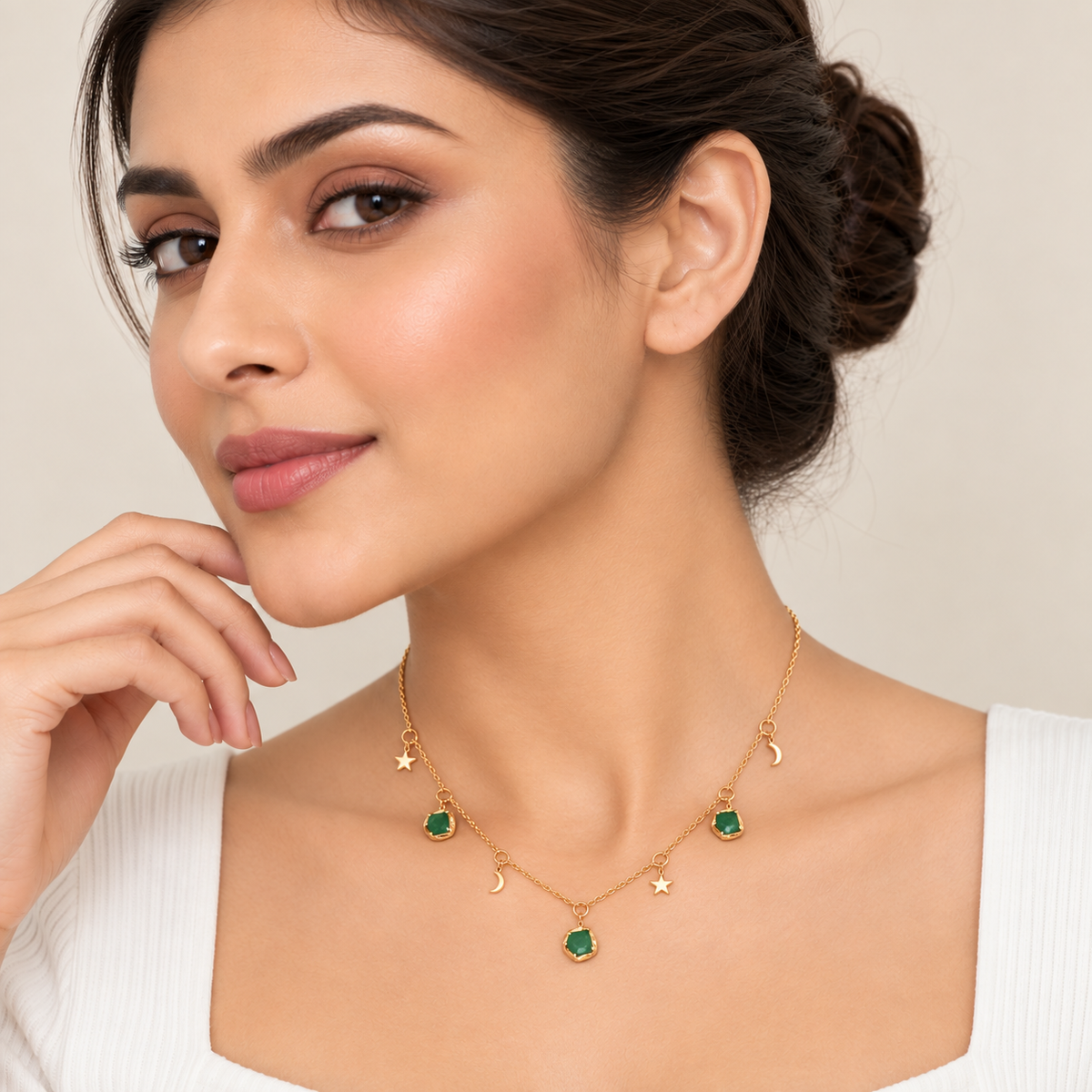 A woman wearing a delicate gold necklace with green gemstone charms shaped like stars, crescent moons, and a central pendant, styled with matching stud earrings against a soft beige background.

