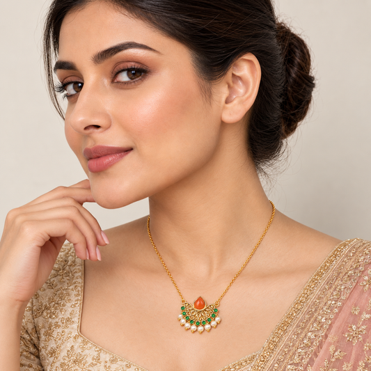 A woman wearing a gold-toned semi-circular pendant necklace with a central orange stone, green accents, and a row of white pearls, shown in a close-up portrait against a soft beige background.

