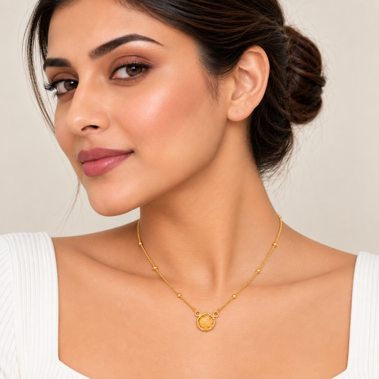 A close-up of a woman wearing a delicate gold chain necklace with a small round pendant, styled with a white ribbed top against a soft beige background.

