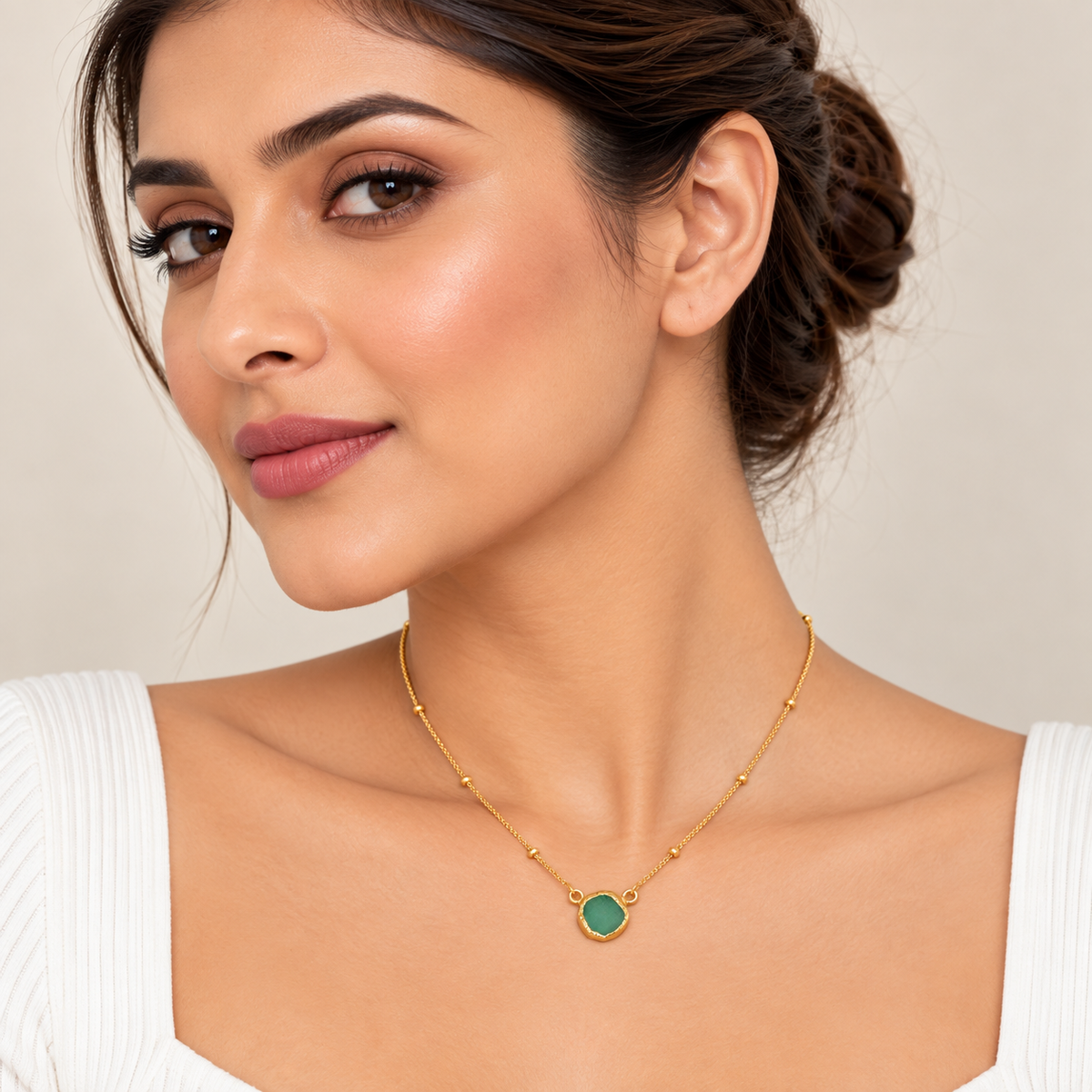 A woman wearing a delicate gold chain necklace with a round green stone pendant, shown in a close-up portrait against a soft beige background.


