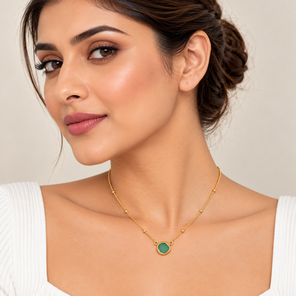 A woman wearing a delicate gold chain necklace with a round green stone pendant, shown in a close-up portrait against a soft beige background.

