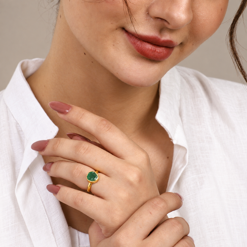 A close-up lifestyle shot of a woman wearing a gold ring with a round green gemstone, styled with a white linen shirt.

