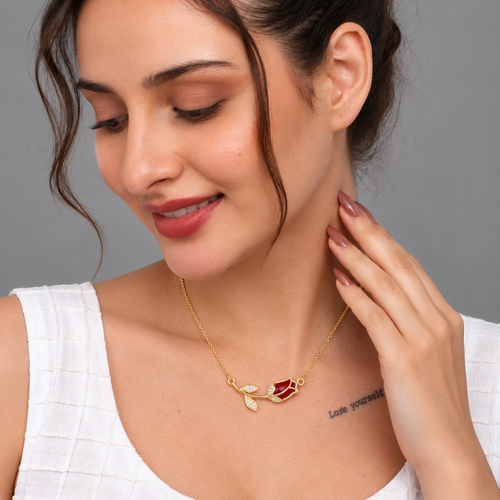 A woman wearing a delicate gold leaf necklace with a red heart-shaped stone and small clear crystals, shown in a close-up portrait against a neutral gray background.

