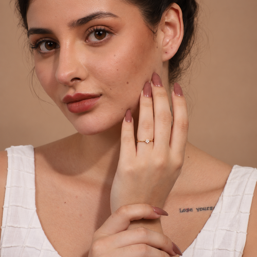 A close-up lifestyle shot of a woman wearing a delicate gold ring with a small clear gemstone on her ring finger. She is wearing a white textured top against a solid warm beige background.

