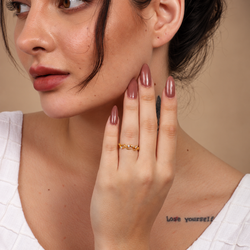 A close-up lifestyle shot of a woman wearing a delicate gold ring with small clear stones on one finger. She has mauve-colored manicured nails and is wearing a white textured top against a warm, neutral background.

