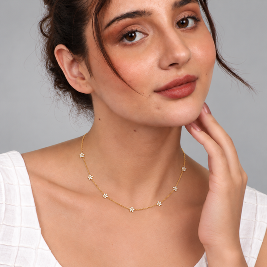 A woman wearing a delicate gold necklace with small daisy-like flower charms, photographed against a plain light gray background.


