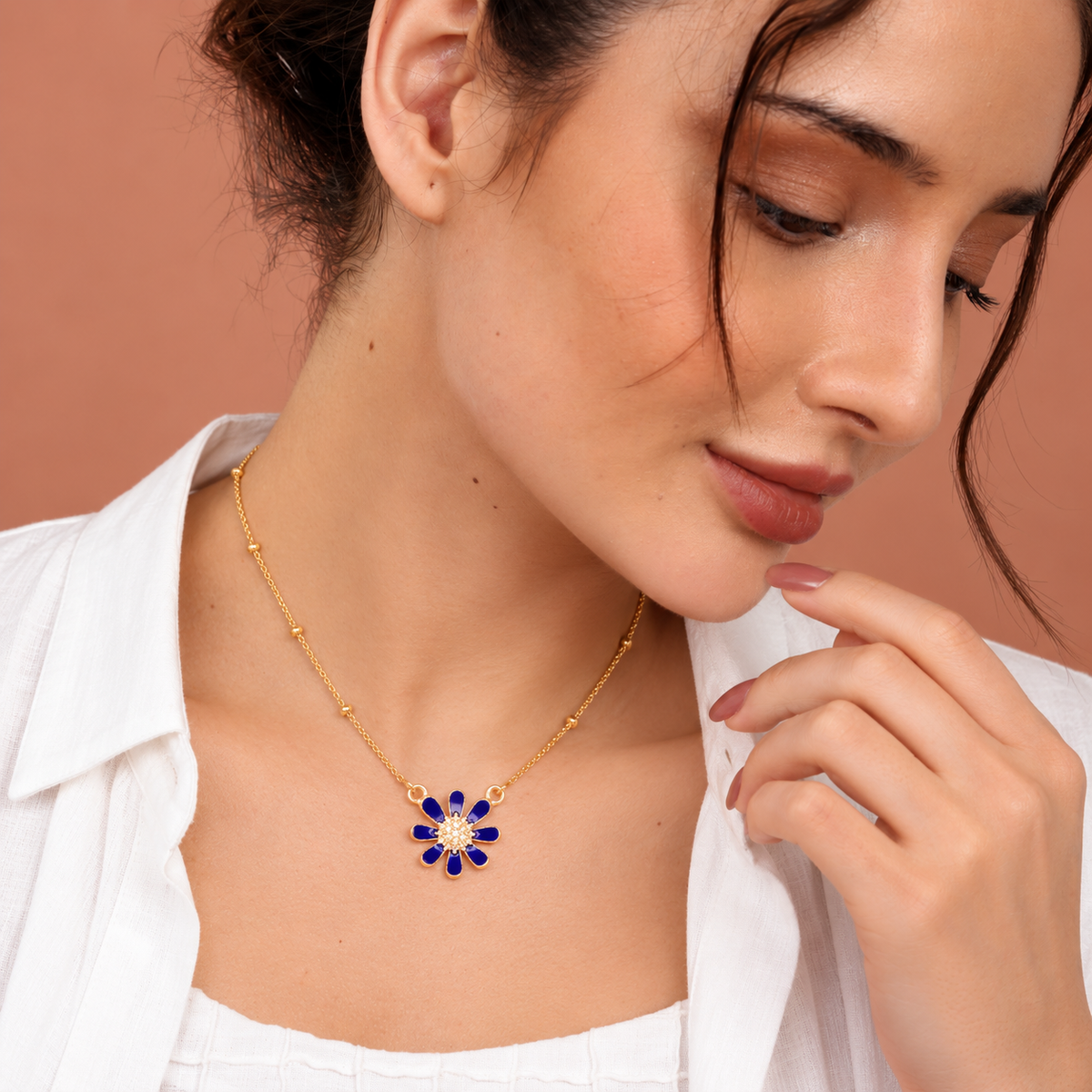A woman wearing a delicate gold chain necklace with a flower-shaped pendant featuring deep blue stones and a small white center, styled with a white collared shirt against a warm peach background.

