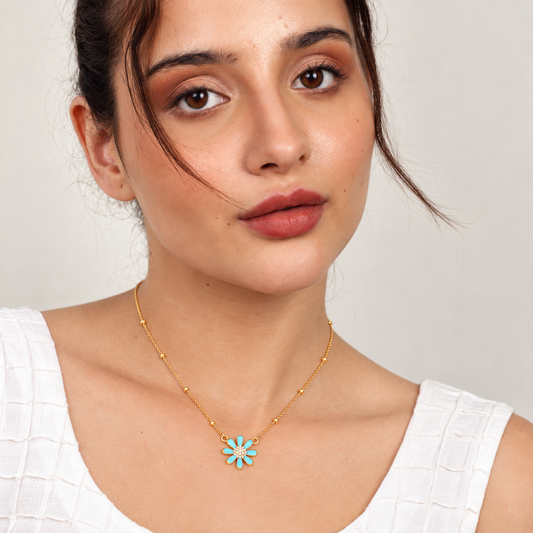 A close-up of a woman wearing a delicate gold chain necklace with a small flower-shaped pendant featuring light blue and white enamel details, paired with a white textured top against a neutral light gray background.

