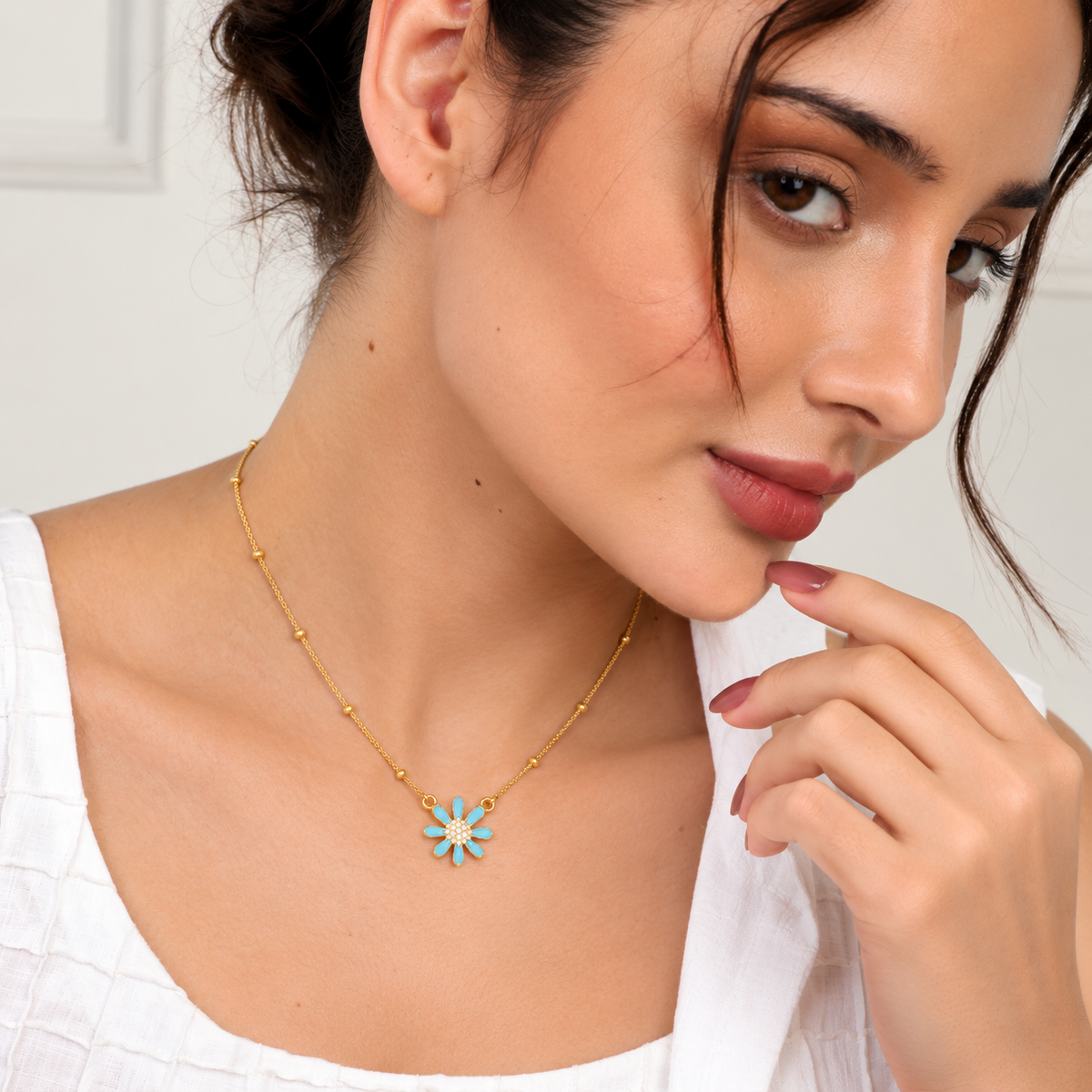 A close-up lifestyle shot of a woman wearing a delicate gold chain necklace with a small flower-shaped pendant featuring light blue and white enamel details.

