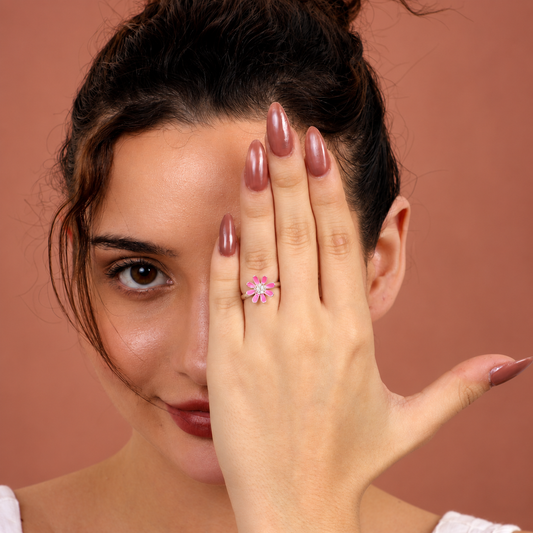 A close-up lifestyle shot of a woman wearing a small pink flower ring on her finger, holding her hand up to show off the ring against a solid warm terracotta background.

