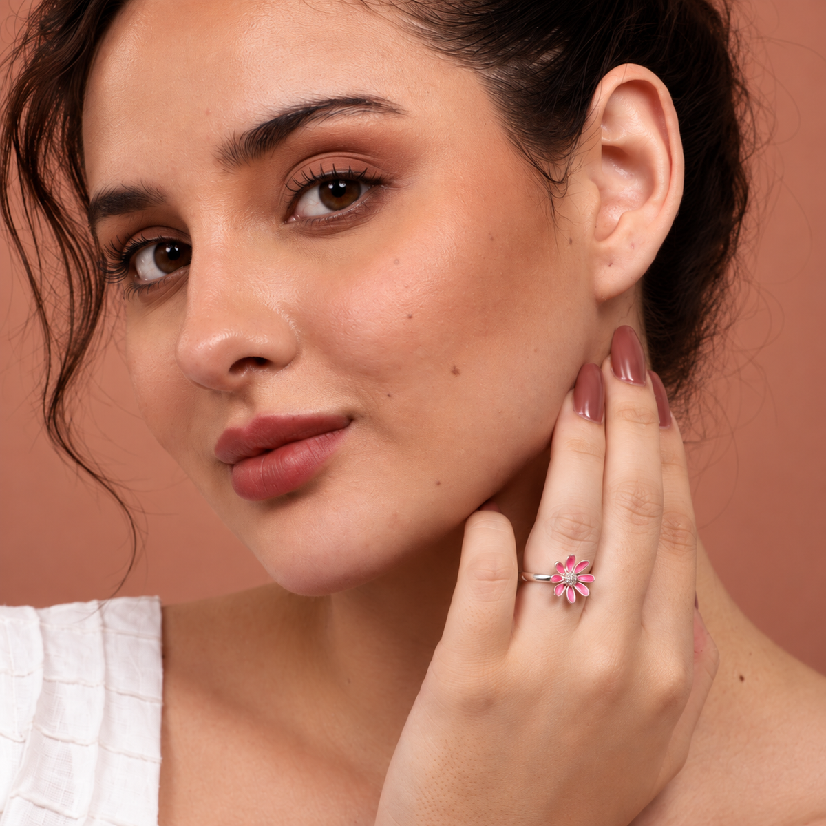 A close-up lifestyle shot of a woman wearing a silver ring shaped like a pink flower, touching her cheek. The background is a solid warm terracotta color.

