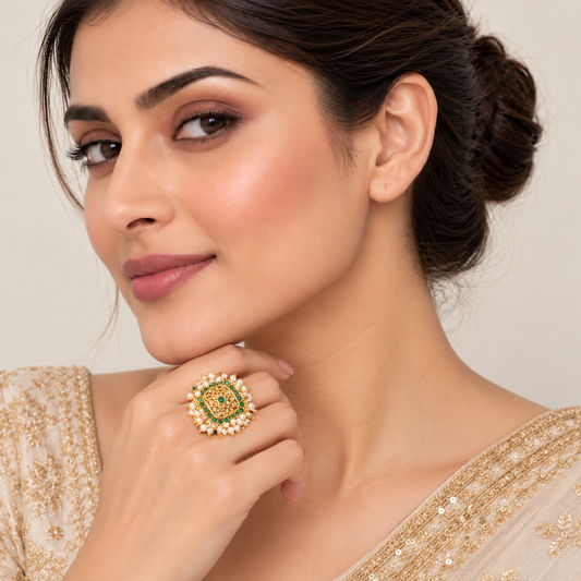 A close-up lifestyle shot of a woman wearing a large, ornate gold ring with a green and red patterned center and white stones, paired with a beige embellished garment.

