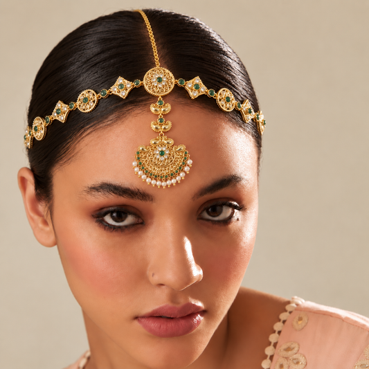 A close-up of a woman wearing an ornate gold maang tikka with green and white stones, set against a warm beige background.

