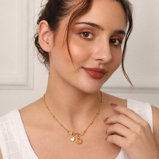 A woman wearing a delicate gold chain necklace with small bead details and three charms: a heart, a crescent moon, and a round medallion. She is wearing a white top against a soft, light-colored paneled wall.

