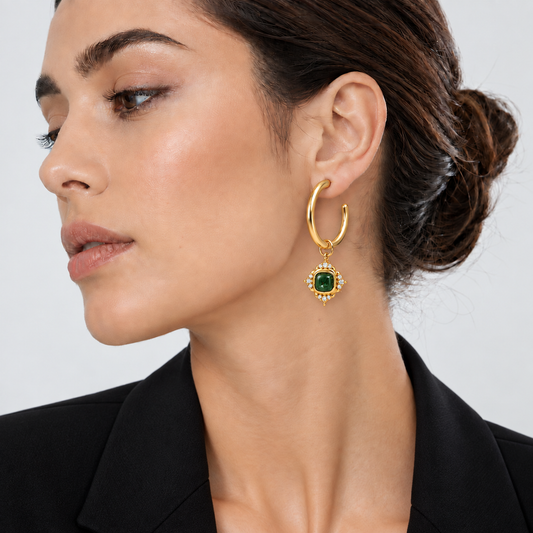 A close-up of a woman wearing a gold huggie hoop earring with a dangling green emerald and gold floral setting, paired with a black blazer against a light gray background.


