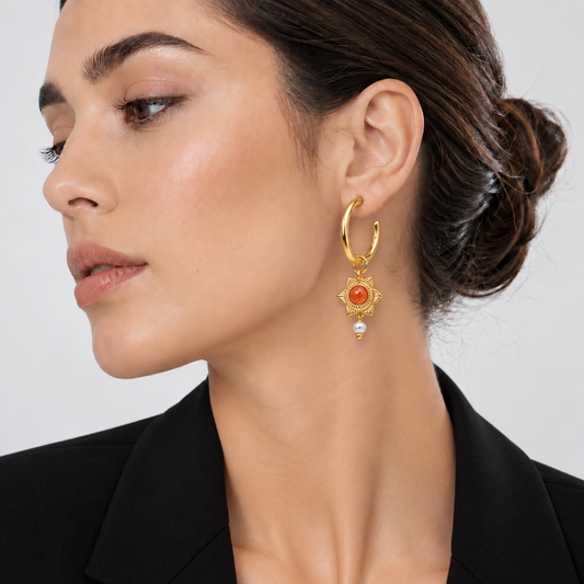 A close-up of a woman wearing a gold huggie hoop earring with a dangling green emerald and gold floral setting, paired with a black blazer against a light gray background.

