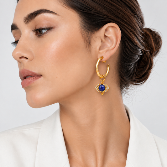 A close-up of a woman wearing a gold huggie hoop earring with a dangling eye-shaped charm featuring a deep blue stone and small clear stones, paired with a white blazer against a light gray background.

