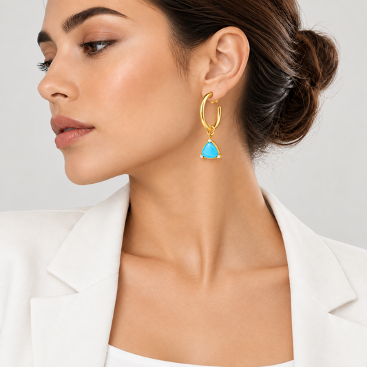 A close-up of a woman wearing a gold hoop earring with a dangling turquoise blue triangle charm, paired with a white blazer against a light gray background.

